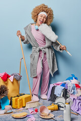 Full length shot of positive housewife with curly hair exclaims loudly holds dusting brush as if...