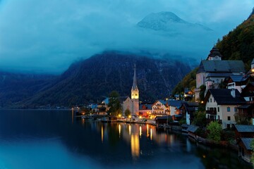 Fototapeta na wymiar View of Hallstatt before dawn with foggy alpine mountains in background & lights of the church & town houses reflected on lake water in twilight, a peaceful lakeside village in Salzkammergut, Austria