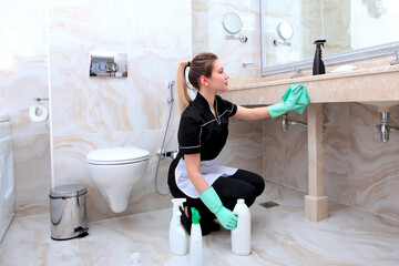 Cleaning in bathroom. A girl in uniform wipes a marble countertop. Cleaning and disinfection of the toilet. The concept of cleanliness . - obrazy, fototapety, plakaty