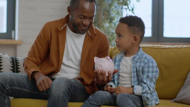 Financial literacy. African american father teaching little son to save money, putting coins into piggybank, free space