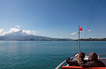 Tourists sitting in the deck of a cruise boat with a Swiss flag waving in the wind on Lake Lucerne...