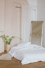 Interior of a stylish bedroom. Bed with white duvet, floor mirror, vase with live flowering branches.