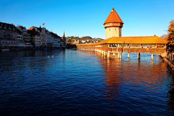 Scenery of Chapel Bridge ( Kapellbrucke ) and Water Tower bathed in warm golden sunlight over Reuss River in Lucerne Old Town, Switzerland, with beautiful reflections on the water under blue clear sky