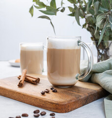 Two cappuccino or latte with milk foam in glasses on a wooden plate with coffee beans, cinnamon and eucalyptus branch close up. The concept of spring breakfast.