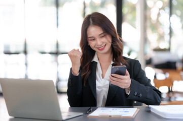 Happy young Asian business woman successful excited raised hands rejoicing with smartphone.