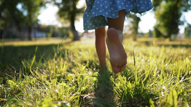 Little cheerful girl with bare feet run along green grass in nature.Foot of child is large on grass in rays of sun.Active childhood outdoors.Healthy lifestyle.Girl child play and run in park in summer