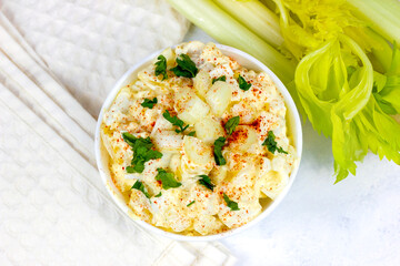 Fresh homemade traditional old-fashioned Amish Potato Salad with potatoes, boiled eggs, creamy dressing and celery in a white bowl on light background top view.