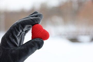 Red knitted heart in female hand in black leather glove on snow park background. Concept of romantic love, Valentine's day, winter weather