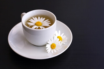 Obraz na płótnie Canvas Chamomile tea in a white cup, daisy flowers on saucer. Soothing and healing herbal drink on dark wooden table