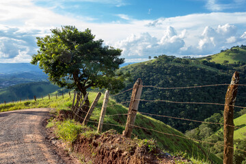 Fence and tree in the foreground with blue sky and hill in the background. Green mountains of Serra da Mantiqueira in the state of Minas Gerais, Brazil