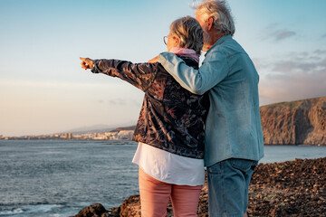 Back view of senior couple standing at sunset light on the rocks at sea looking at the horizon over...