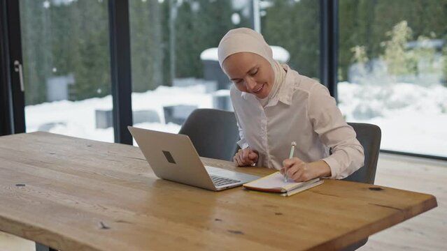 Cute female in hijab is taking notes at the table