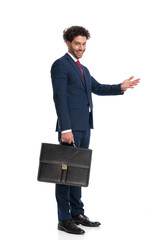 elegant businessman in suit inviting to side while smiling and holding suitcase