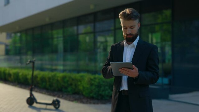Bearded businessman in formal suit , using the tablet for working purposes standing outside near office centre. Male manager texting, swiping apps using digital tablet.