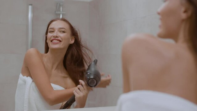 Young carefree woman drying her wet hair after shower, looking at bathroom mirror at home, tracking shot, free space