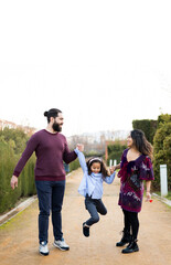 Obraz premium The interracial family is active and enjoys a day at the park. Successful adoption. The parents swing the daughter with their arms while they watch. Concept of multiethnic or interracial family.