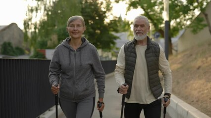 Portrait of the senior couple with trekking poles walking in the street. Practicing scandinavian walking outdoors. Aging youthfully. Elderly people and sport concept.