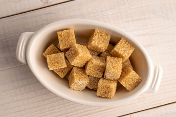 Fototapeta na wymiar Several brown sugar cubes in a white ceramic bowl, close-up, on a white wooden table.