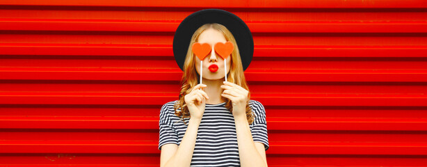 Portrait of beautiful young woman covering her eyes with red heart shaped lollipop wearing black...