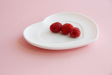 ripe raspberries on a white plate in the shape of a heart on a pink background. raspberry berry on a plate