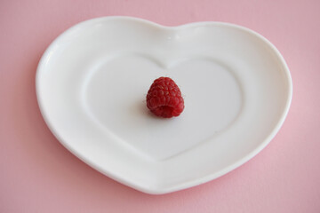 ripe raspberries on a white plate in the shape of a heart on a pink background. raspberry berry on a plate
