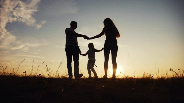 happy family mom dad and daughter play round dance silhouette at sunset fun. people in park kid dream concept. happy family parents with little kid child daughter play in park on grass holding hands
