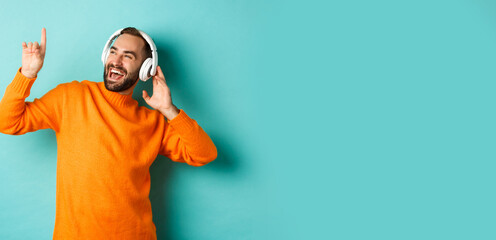Happy adult man in orange sweater, looking up and listening music in headphones, standing over blue background