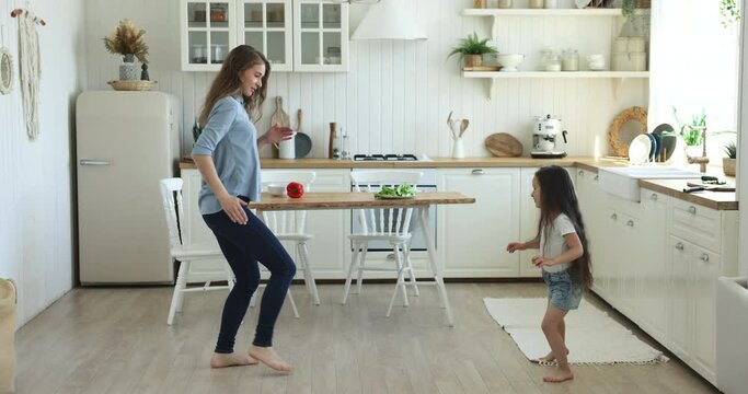 Pretty woman dance barefoot with cute little 5s daughter, moving to favourite music, hold hands, spinning, having fun in warm modern kitchen on weekend at home. Hobby, leisure with kid, active pastime