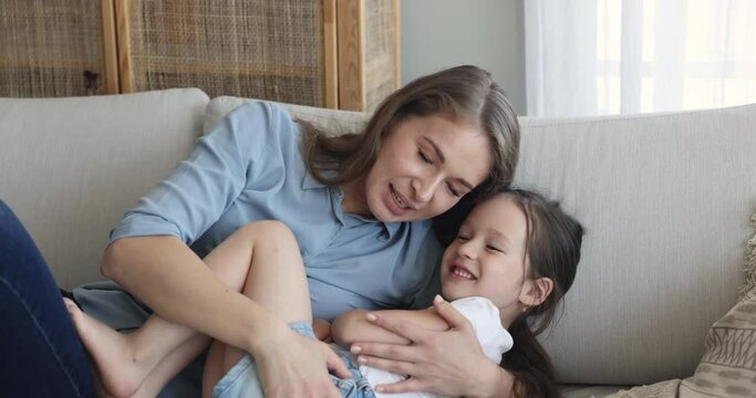 Close up loving young mother or babysitter play with cute little preschooler daughter girl, lying together on cozy couch having fun feel happy. Mum and kid enjoy carefree leisure at home. Family bond