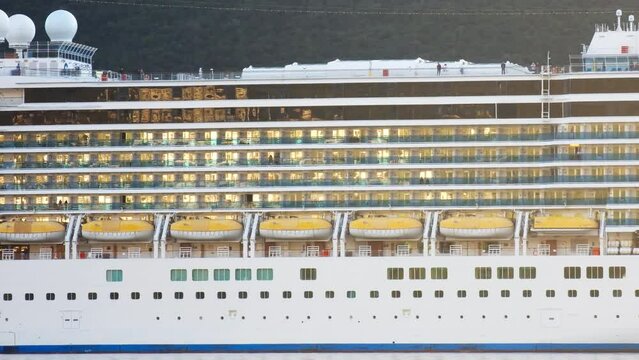 Cruise ship - balconies on the decks, lifeboats and portholes, on port or starboard side of a luxury boat, light in windows. Wealthy tourists spend summer vacation traveling in rooms and cabins. 