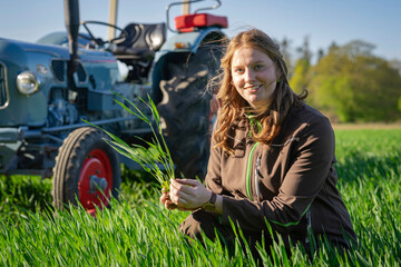 Landwirtin überprüft das junge Getreide auf einen Getreideacker, sie hält eine Pflanze in ihrer Hand, im Hintergrund ein Oldtimertracktor. © Countrypixel