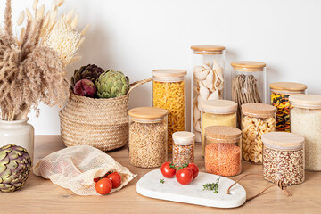 Assortment of grains, cereals and pasta in glass jars and kitchen utensils on wooden table. Healthy...