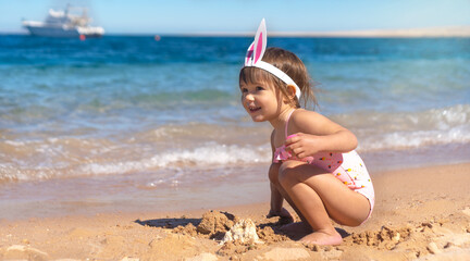 Little girl in swimsuit and bunny hairband sitting on the sandy beach. Happy Easter holidays...