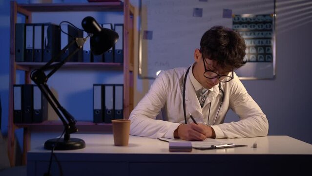 Young arab doctor sitting at table and writing on a document report in hospital office. Medical healthcare staff and doctor service concept.