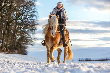 A young equestrian teenage girl rides on her haflinger horse through the snow in the evening during...