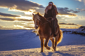 A young equestrian teenage girl rides on her haflinger horse through the snow in the evening during sundown in front of a snowy rural winter landscape