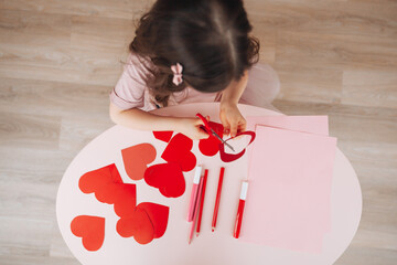 a little girl makes Valentine's Day cards using colored paper, scissors and pencil, sitting at a table in a cozy room.