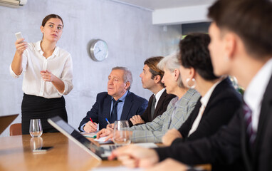 Confident interested young businesswoman doing presentation to colleagues during work meeting in office, using remote control to switch slides