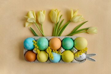 colorful painted Easter eggs in a paper egg container with tulips on a beige background. Top view