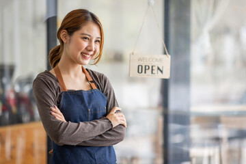 Shot of smiling young cafe show owner Asian woman standing with arms crossed and Open sign on the glass door. Portrait of asian tan woman barista cafe SME entrepreneur seller business concept.