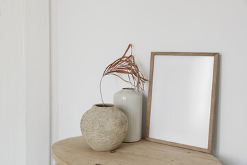 Blank wooden picture frame mockup on old round console table. Dry palm leaf in textured vase. White wall background. Empty copyspace. Elegant boho interior. Living room, artistic atelier display.