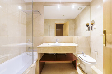 Bathroom with cream-colored marble furniture with an elongated bathtub and a mirror embedded in the wall and a metal shelf inside and a glass partition