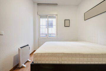 Bedroom mattress without bedding, an electric radiator and window with metal bars
