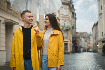 Lovely young couple with umbrella walking under rain on city street