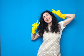 young girl cleaner in uniform and gloves for cleaning shows a gesture of peace and poses on a blue background
