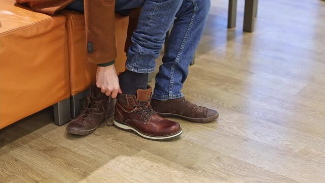 Young handsome caucasian brunette in a brown coat puts on a new shoe for trying on, sitting on a leather pouffe in a shoe store, close-up side view. The concept of offline shopping, men's lifestyle.