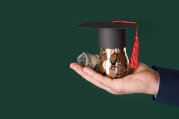 Man holding glass jar of coins, dollar banknotes and graduation cap against green background,...