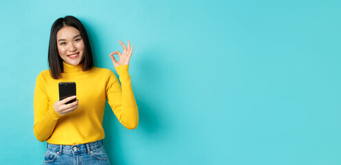 E-commerce and online shopping concept. Portrait of asian woman showing OK sign and using mobile phone, praise app, standing over blue background