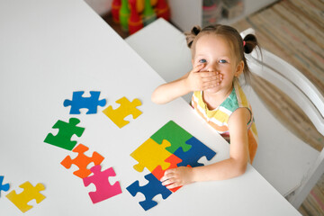 A surprised kid is sitting at a table collecting colorful puzzle pieces. Preschool education of...