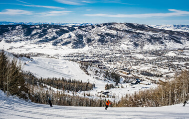 View of Colorado, USA, ski slope descending to ski resort village in winter with three figures of snowboarders going down the slope; distant mountains and blue sky in background